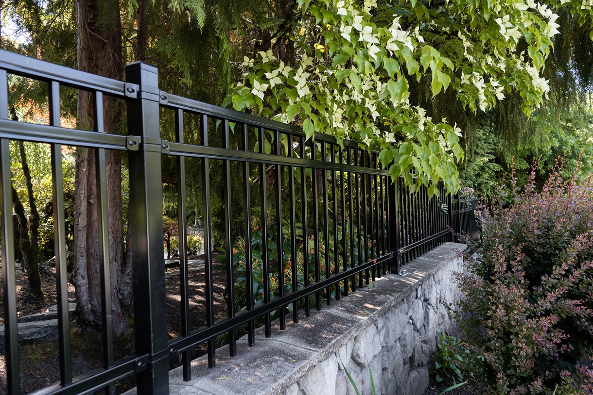 A black metal fence with vertical bars and square posts, mounted on a stone retaining wall in a landscaped garden