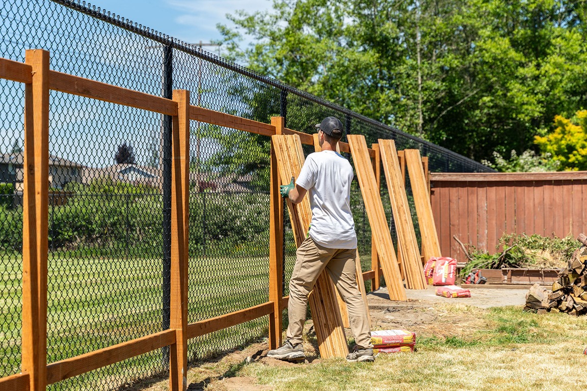 worker in a white shirt and cap installs vertical wooden planks onto a wooden frame in front of a black chain-link fence