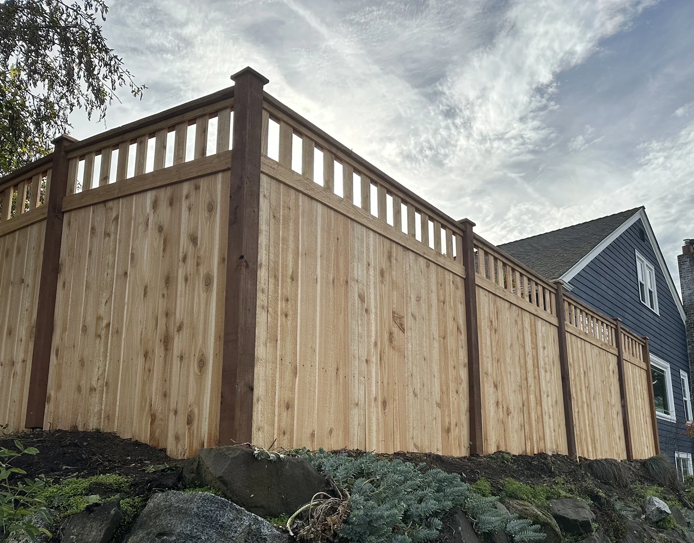 Tall wooden privacy fence with decorative slats atop a rocky garden beside a blue house.