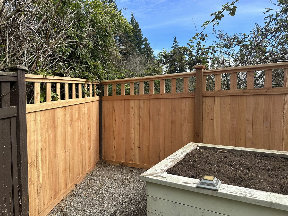 Corner of a wooden fence with gravel ground and a white planter box filled with soil.