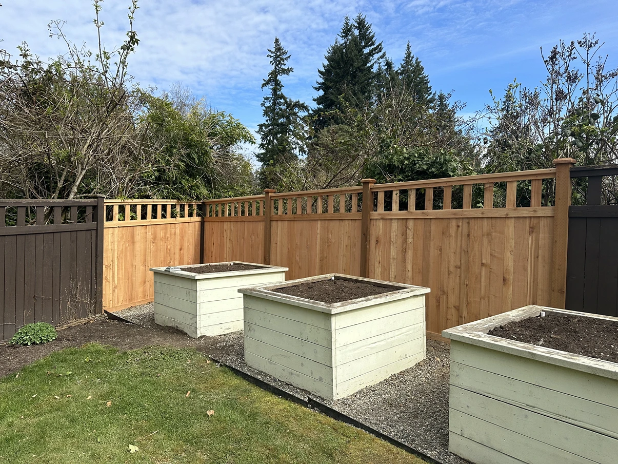 Three empty raised garden beds in a backyard with wooden fence and trees under a cloudy sky.