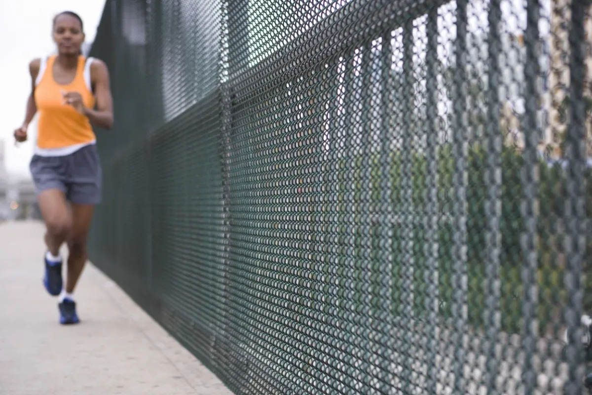 Active woman in orange tank top and blue shorts jogging on a city sidewalk next to a metal fence.