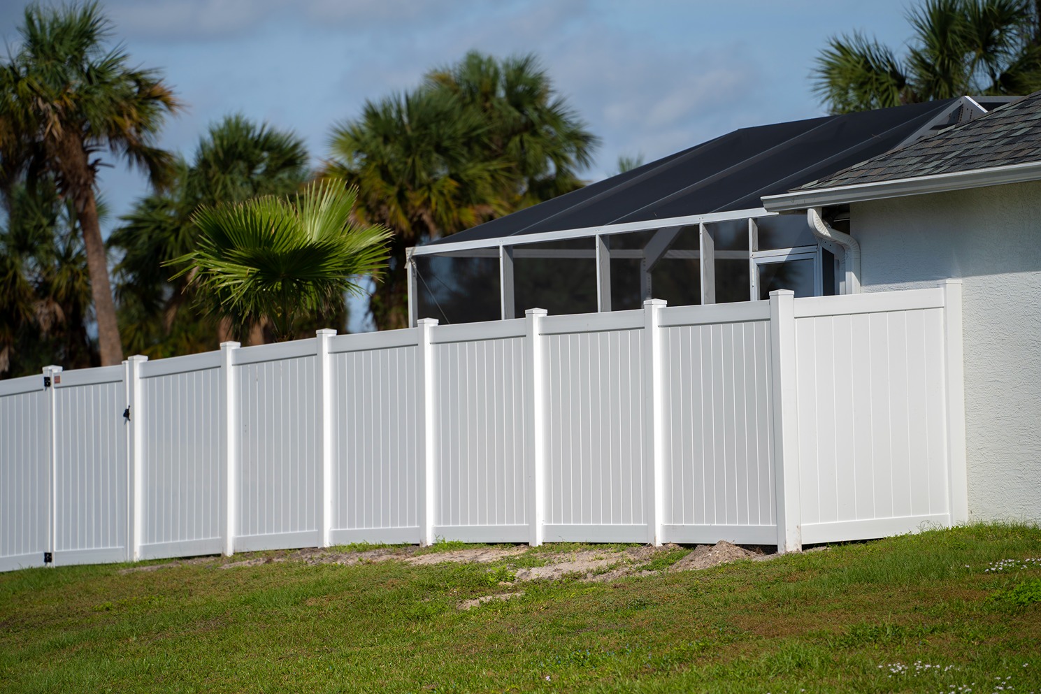 White vinyl privacy fence along a grassy yard with palm trees and a screened porch in background.