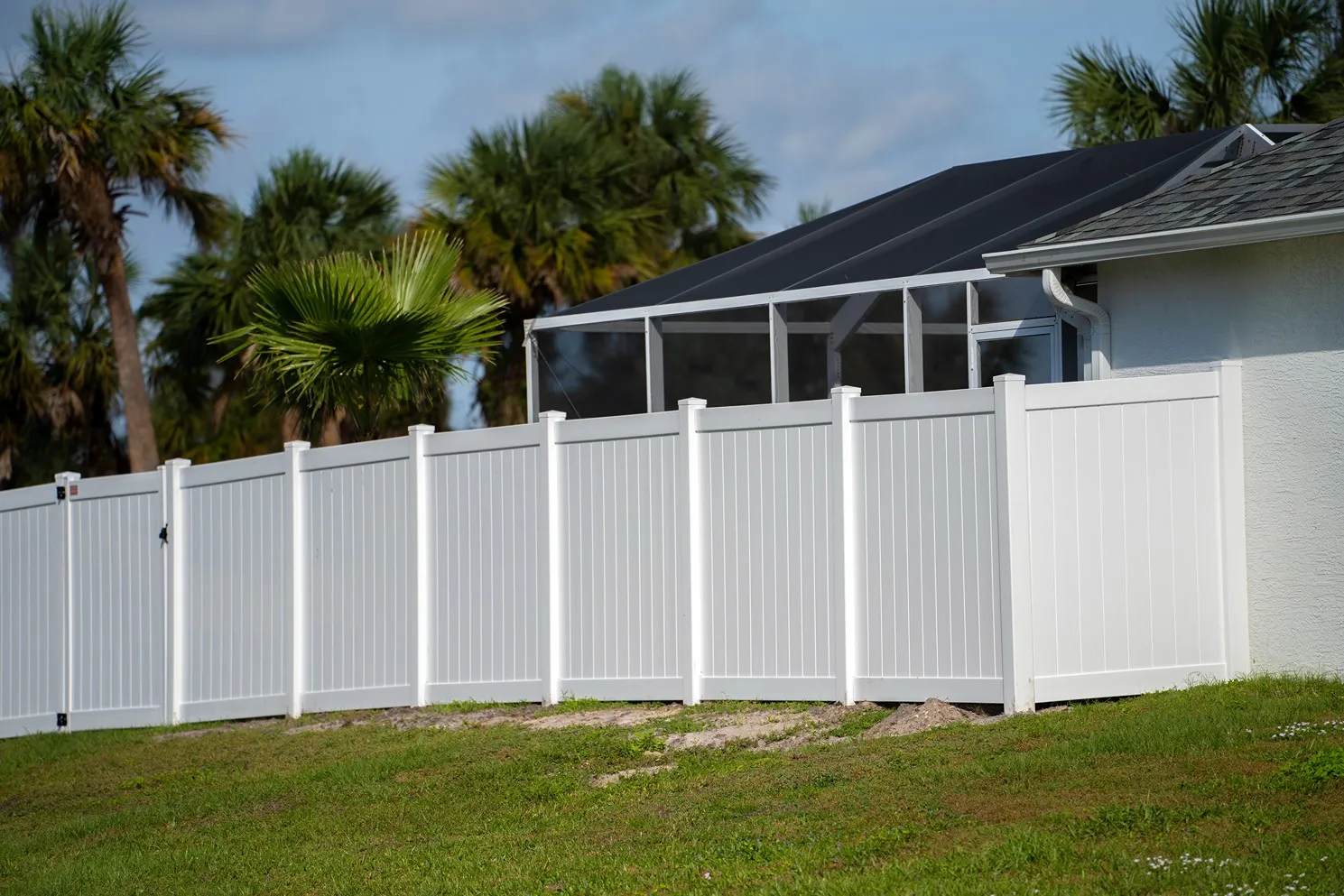 White vinyl privacy fence along a grassy yard with palm trees and a screened porch in background.