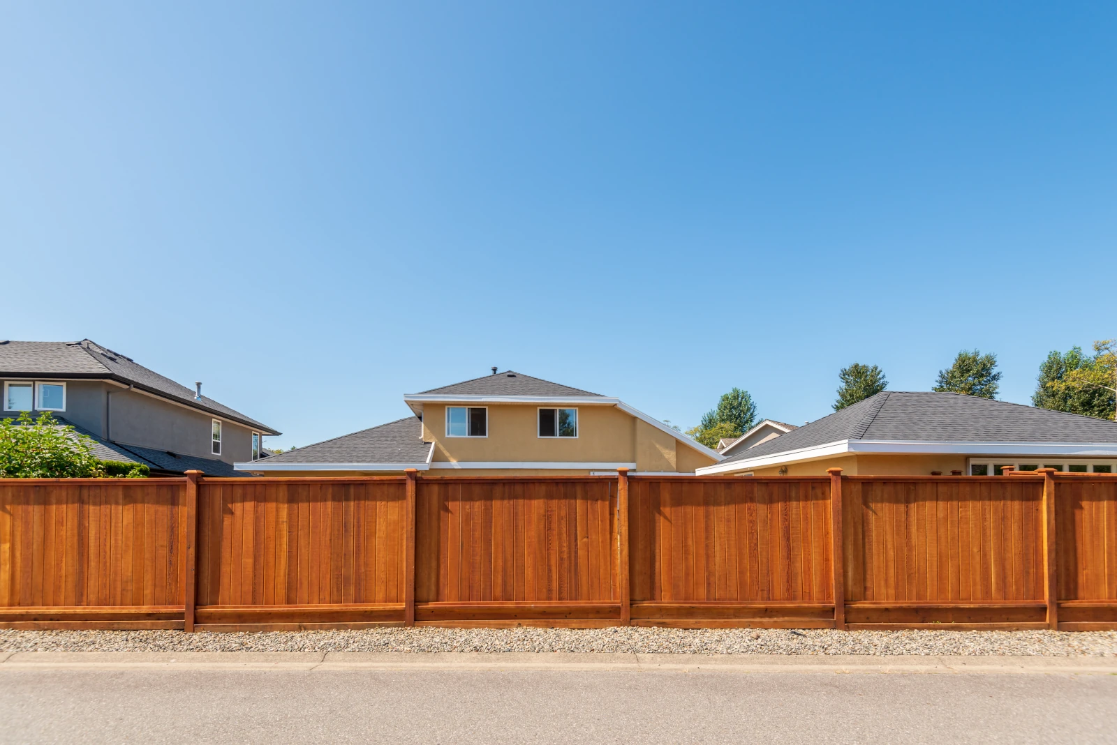 Tall wooden privacy fence with vertical panels in front of suburban houses under clear blue sky.