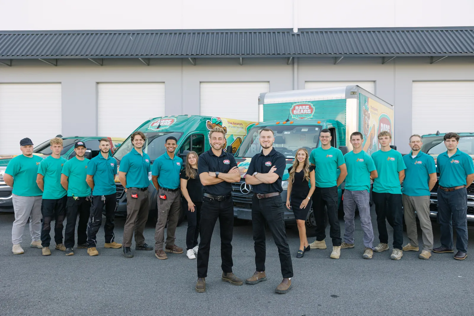 Rare Bears Fencing crew in teal uniforms posed in a V formation with two team leads in front, backed by branded vans and a box truck at the shop