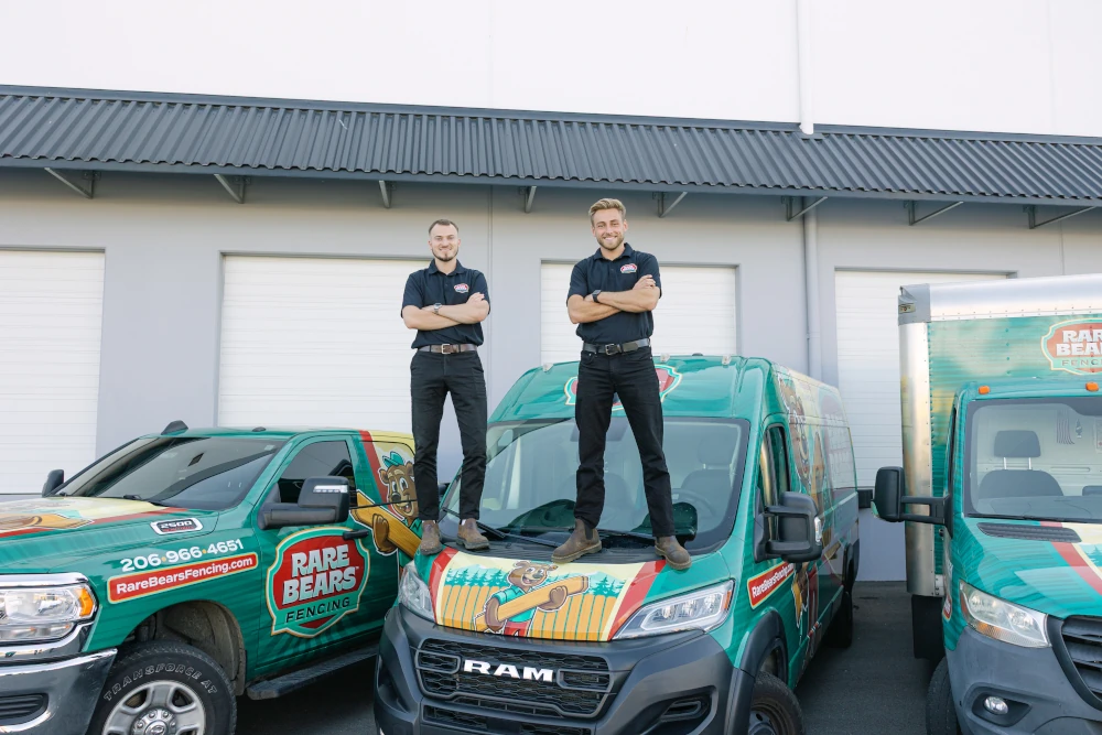 Two Rare Bears Fencing owners standing on the hood of a branded work van with the company truck fleet at the shop