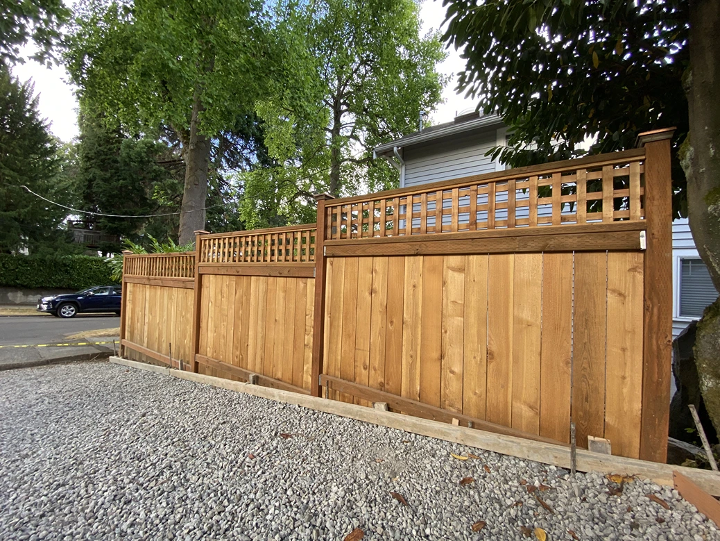 Three wooden fence panels with lattice tops are installed on gravel ground, with tall green trees and a gray house in the background.