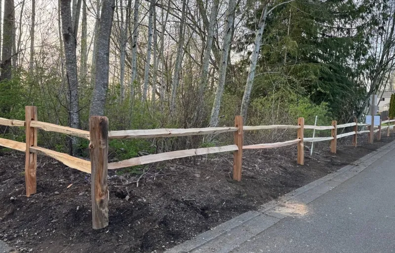 Two-rail cedar fence marking a property border along a planted bed at the edge of a wooded lot