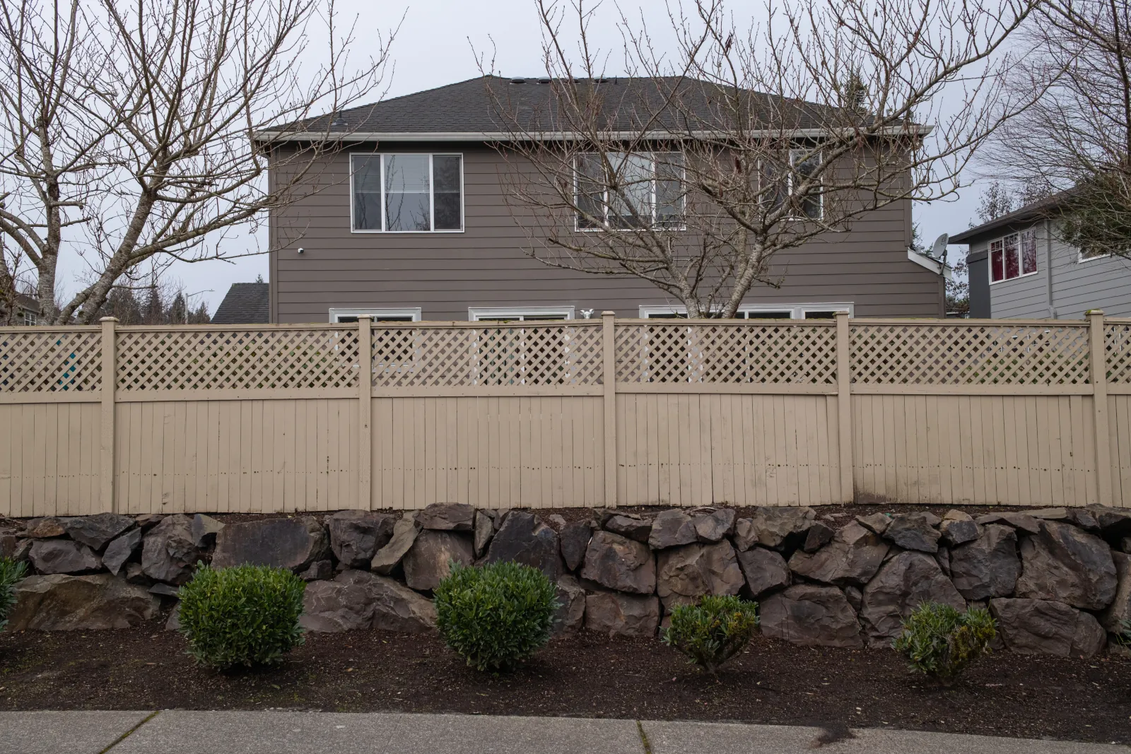 Aging tan-painted wood fence with diagonal lattice top along a rock retaining wall