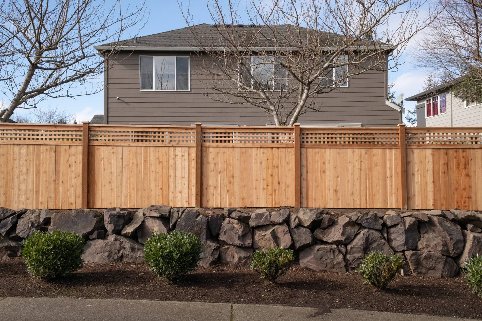 Aging tan-painted wood fence with diagonal lattice top along a rock retaining wall