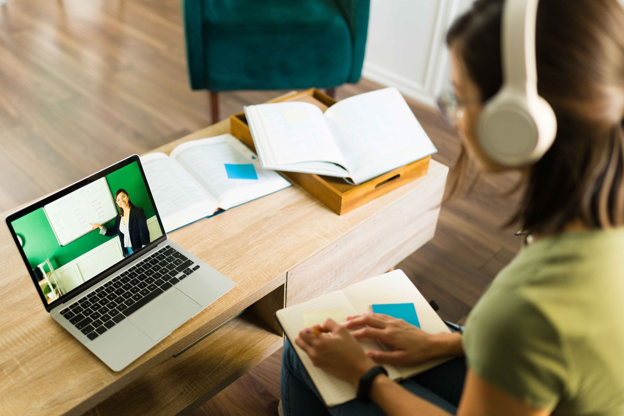 Student attending an online class from home on a laptop, watching a teacher explain lessons while taking notes