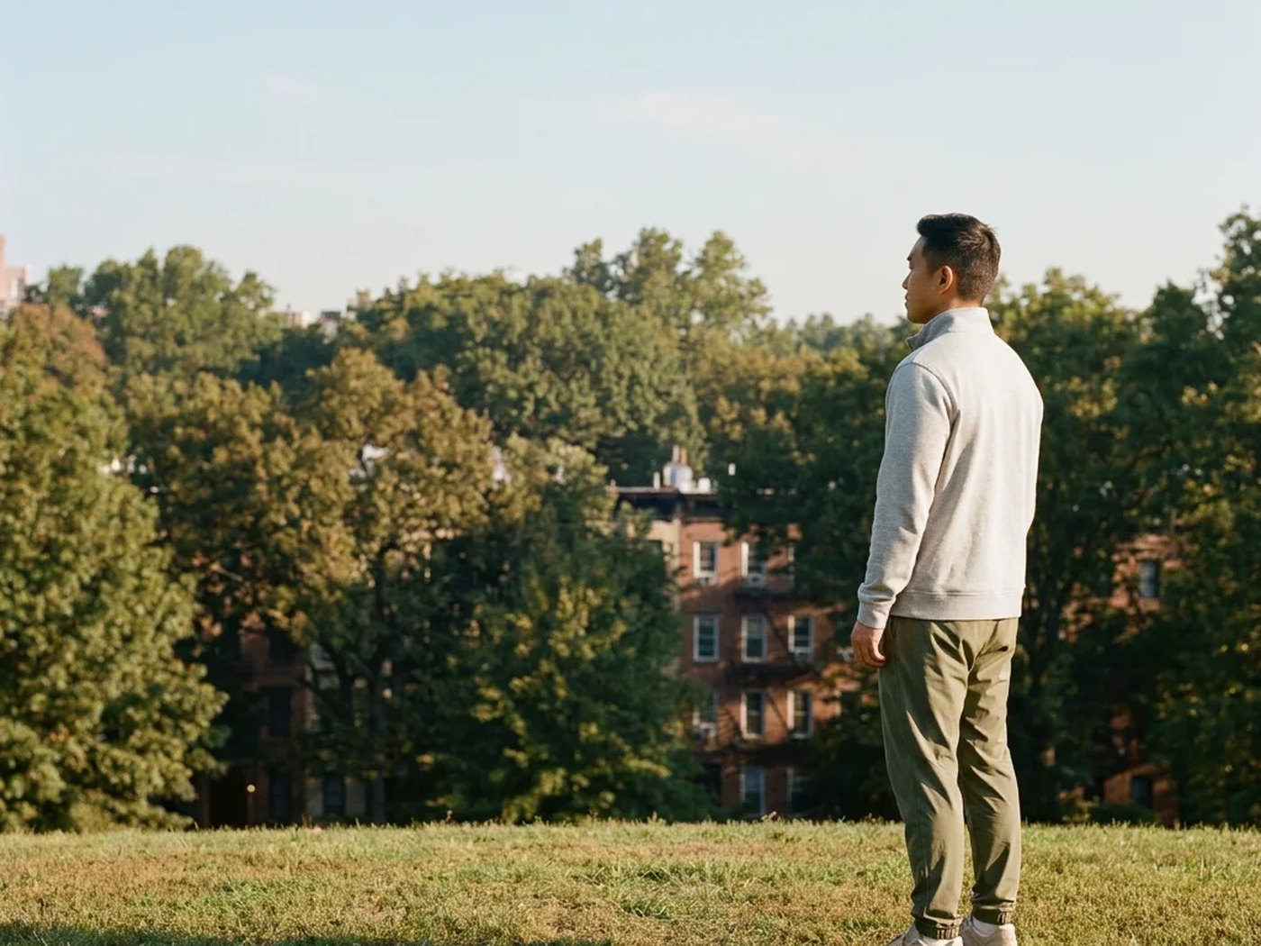 A man standing still on a lawn at the edge of a park, brownstones behind the trees