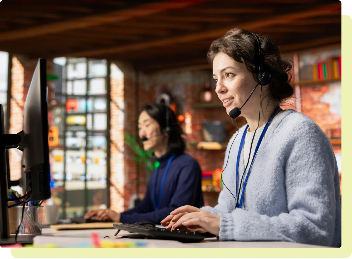 Two customer service representatives wearing headsets working on computers in a bright office.