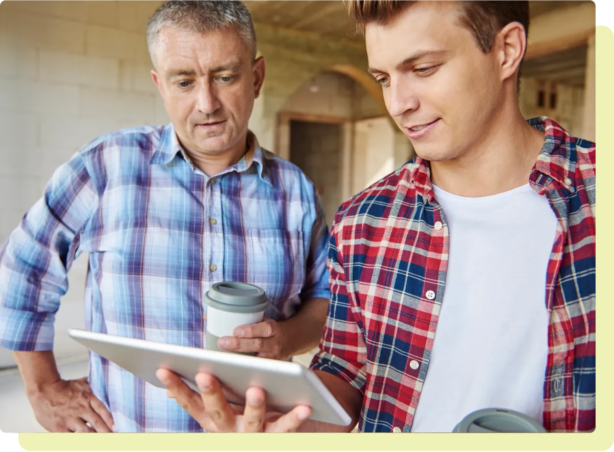 Two men in plaid shirts holding coffee cups looking at a tablet inside an unfinished building.