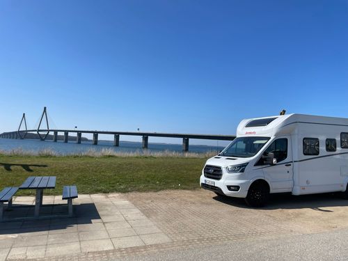 Ein weißes Wohnmobil, das an einem klaren, sonnigen Tag neben einer Picknickbank in der Nähe einer großen Brücke über das Wasser geparkt ist.