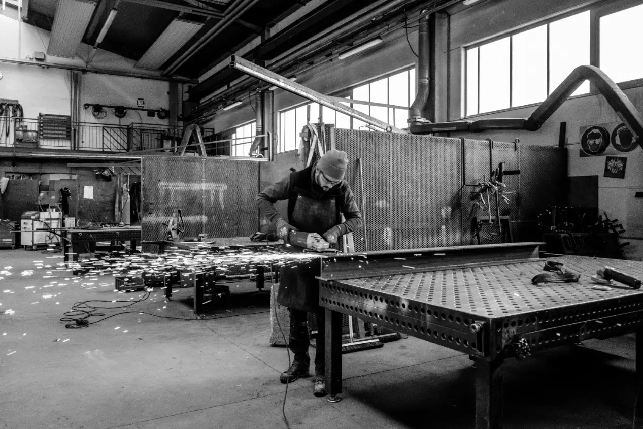 Schwarz-weiss-Aufnahme einer grossen Werkstatt, in der ein Arbeiter mit Mütze und Schutzkleidung an einem Metallstück mit einer Flex arbeitet. Es fliegen zahlreiche Funken, die den Bearbeitungsprozess zeigen. Im Hintergrund sind Werkzeuge, Maschinen und grosse Fenster sichtbar; verwendet für das Produkt „Für Schwärzer Mettal Olang Pustertal“.
