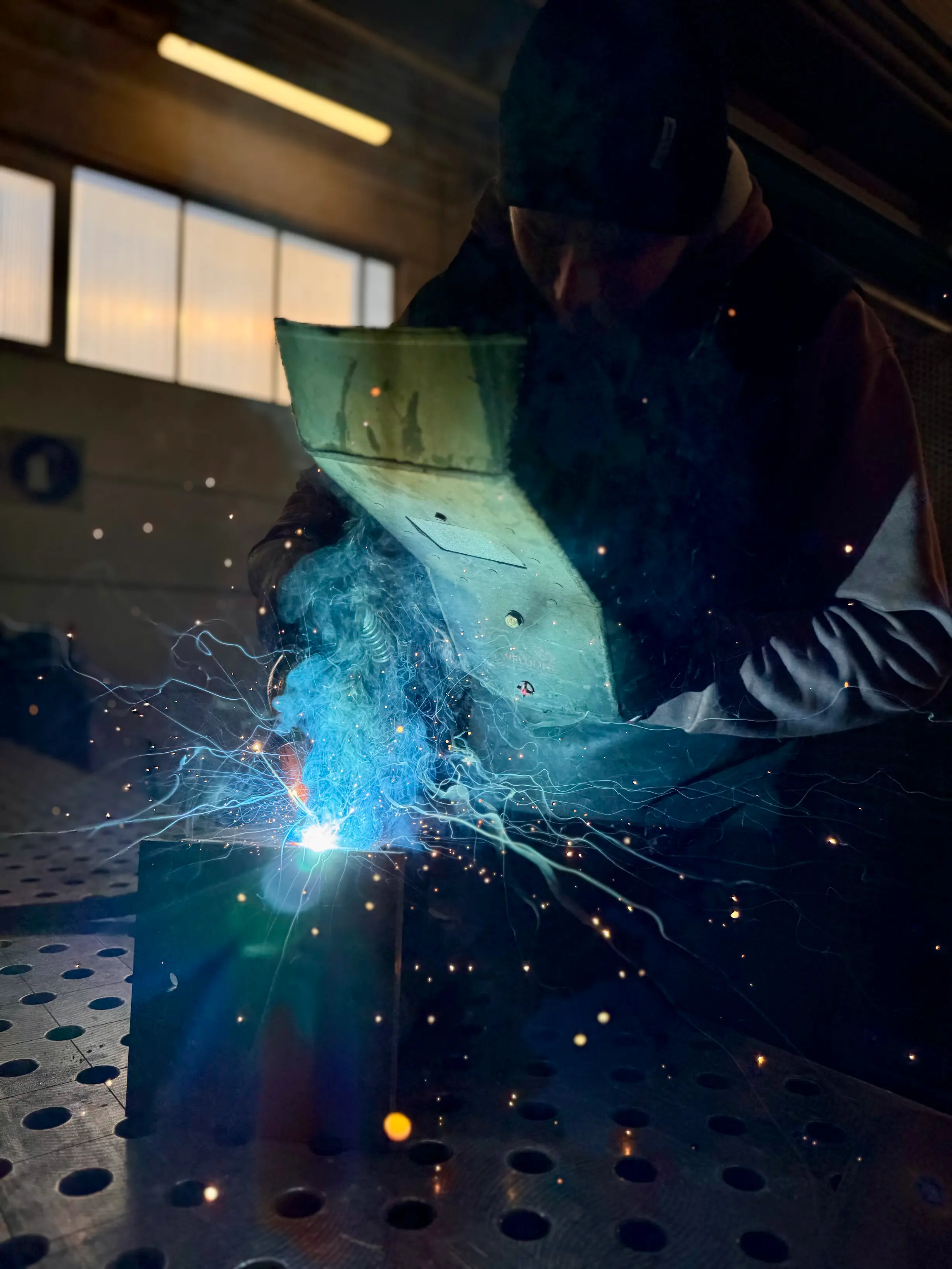 Schweissarbeiten in einer Metallwerkstatt in Olang, Pustertal: Eine Person führt präzise Schweissarbeiten an einem Metallstück durch, mit sichtbaren Funken und Lichtbogen. Im Vordergrund liegt eine gelochte Metallarbeitsplatte. Die Szene zeigt das kunstvolle Handwerk der Metallbearbeitung und Schmiedekunst im Pustertal.