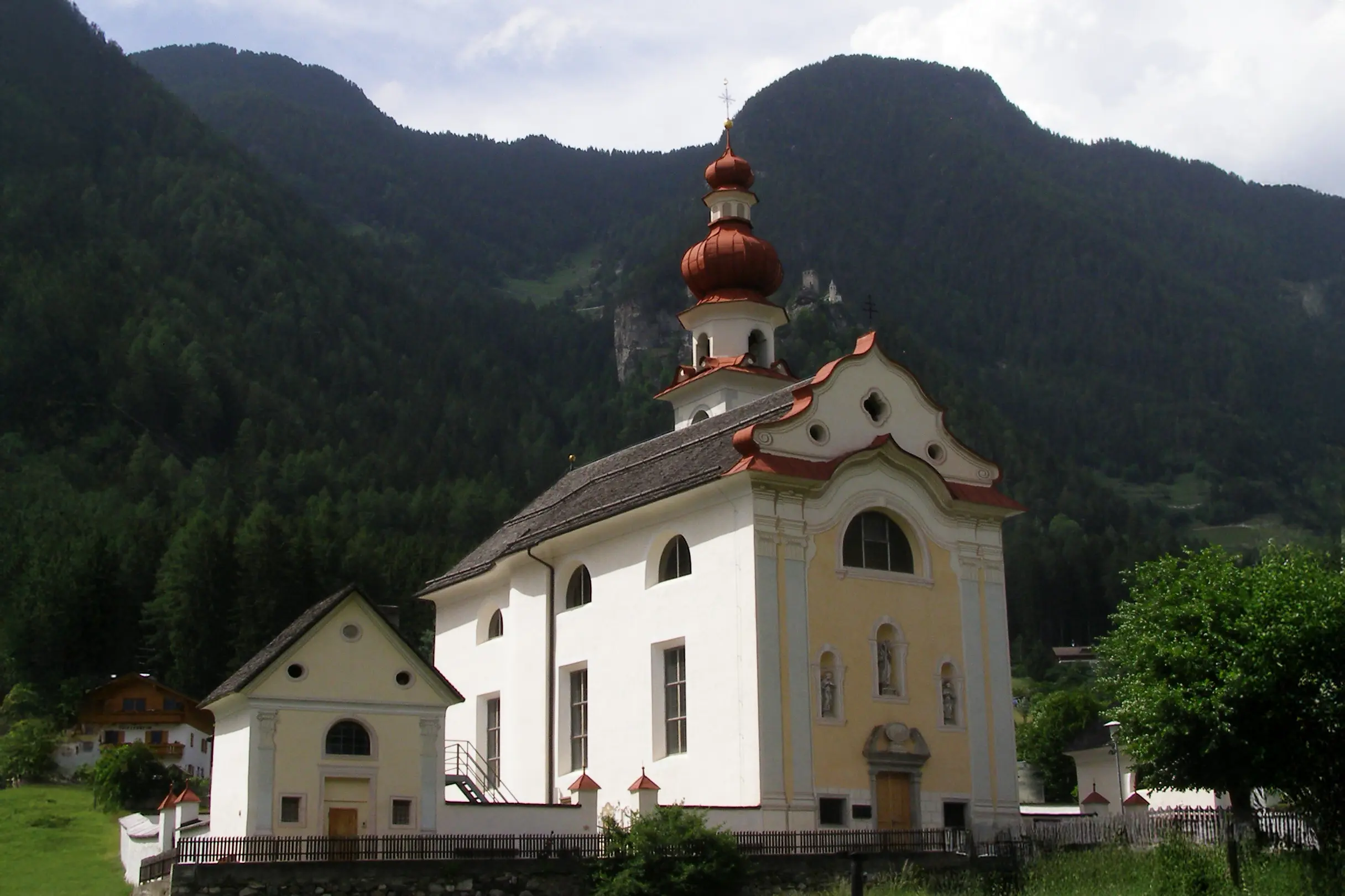Eine weiße Kirche mit rotem Zwiebelturm vor bewaldeten Bergen unter bewölktem Himmel.