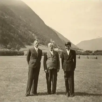Schwarz-weiß-Foto von drei Männern in Anzügen, die auf einer Wiese vor bewaldeten Bergen stehen.