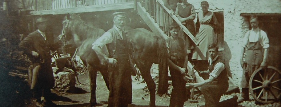 Schwarzweiß-Foto einer Gruppe von Menschen mit einem Pferd vor einem Haus, zwei Männer arbeiten am Huf des Pferdes, andere stehen oder sitzen in der Nähe.