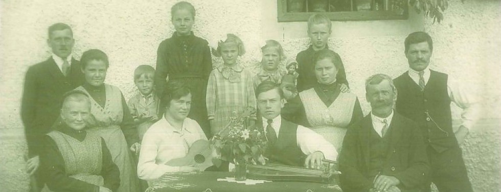 Schwarz-weiß-Familienporträt von 14 Personen, darunter Männer, Frauen und Kinder, einige sitzen um einen Tisch mit Blumen und Musikinstrumenten.