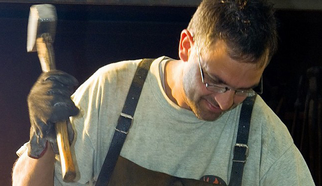 Ein Mann mit Schutzbrille und Arbeitshandschuhen hält einen Hammer in der rechten Hand und blickt konzentriert nach unten. Er trägt ein helles