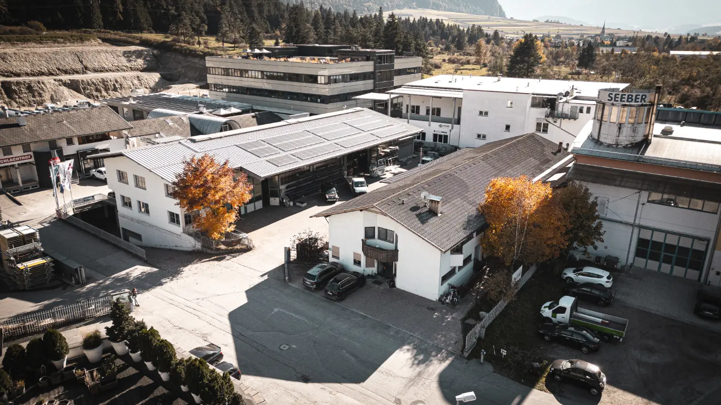 Luftaufnahme eines Industriegebiets mit weißen Gebäuden, herbstlichen Bäumen und mehreren geparkten Autos.