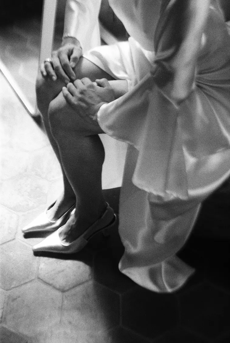 Mariée assise, les mains posées sur sa jambe et ses escarpins visibles pendant les préparatifs à la ferme de Gy en Savoie.