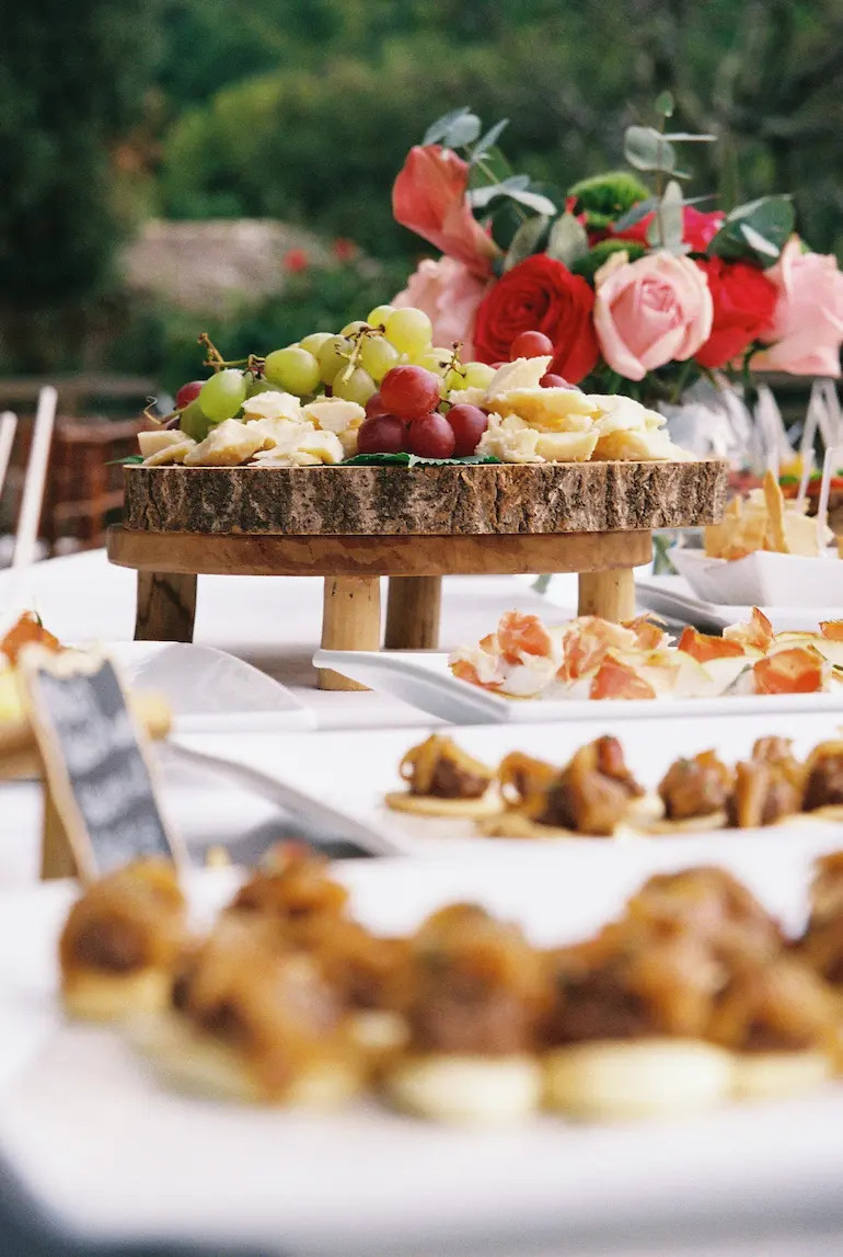 Buffet de mariage en plein air avec fromages, raisins et compositions florales rouges et roses.