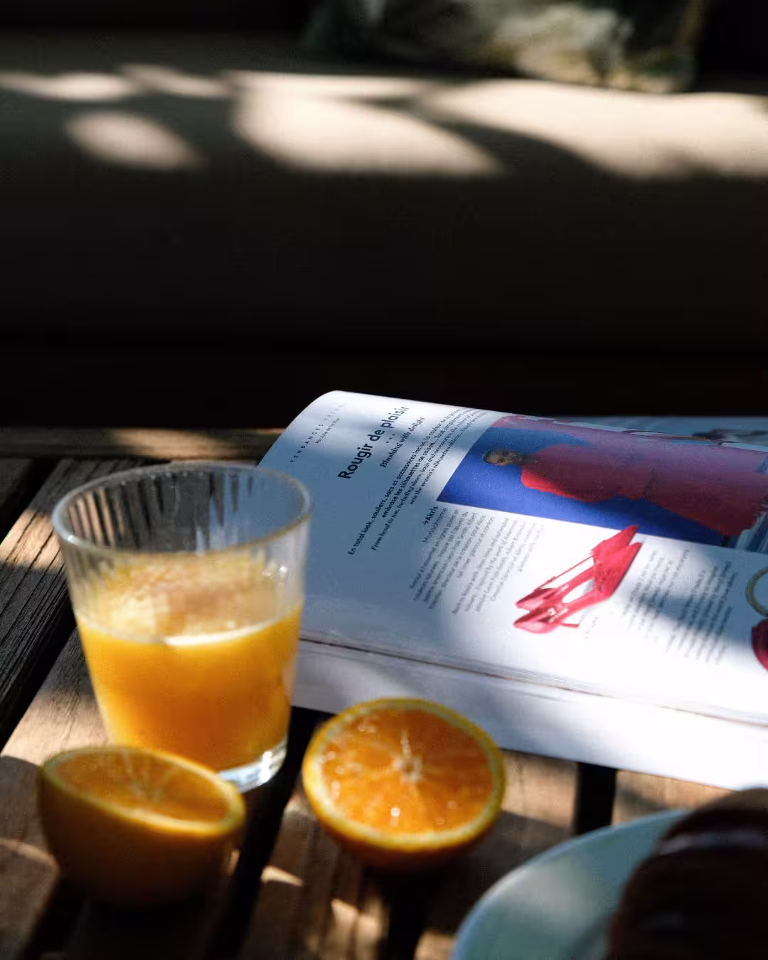 Petit-déjeuner au soleil avec jus d’orange frais et magazine posé sur une table en bois.