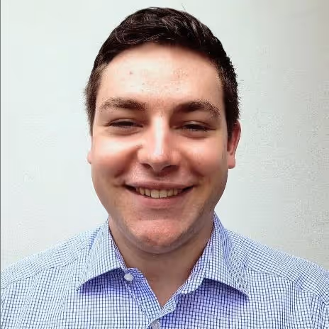 Smiling young man with short dark hair wearing a blue checkered shirt against a plain light background.