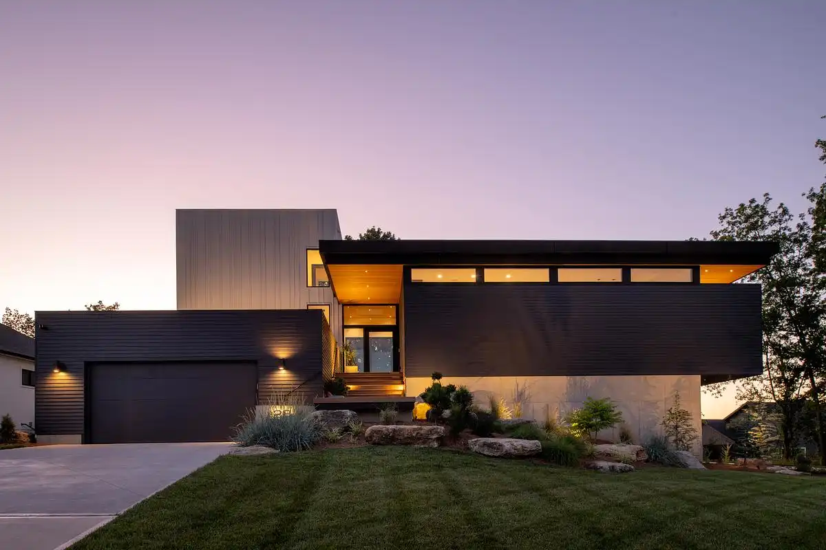 Modern lake house at dusk, its low horizontal roof and lit interior seen across a lawn