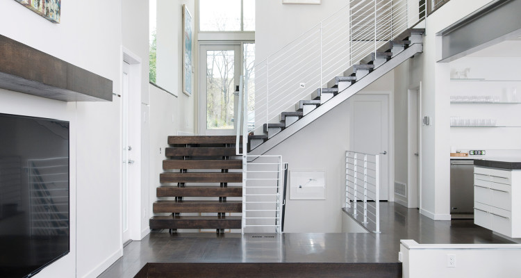 Open stair with wood treads rising through a white double-height interior, with tall windows and a kitchen beyond