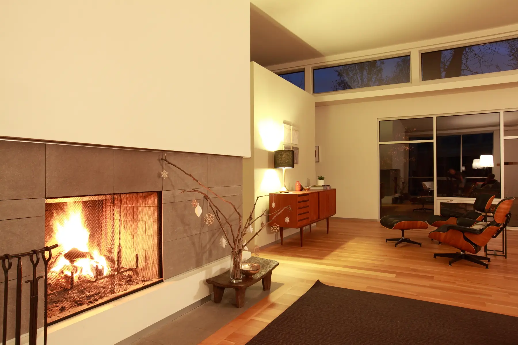 Fire burning in a gray-tiled fireplace, with a wood credenza, lounge chairs and tall windows beyond in evening light