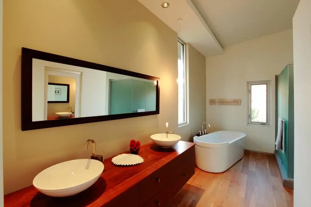 Bathroom with a freestanding tub, two vessel sinks on a wood vanity, a long dark-framed mirror and a green glass shower