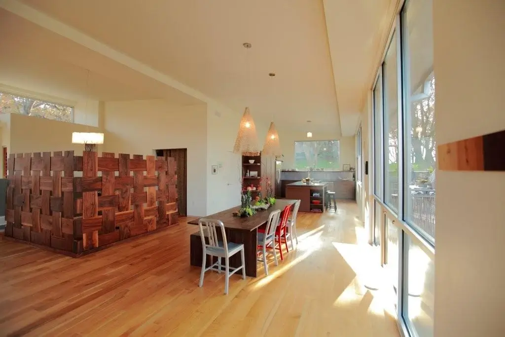 Sunlit dining area along a wall of floor-to-ceiling glass, with a woven wood screen wall, pendant lights and the kitchen beyond