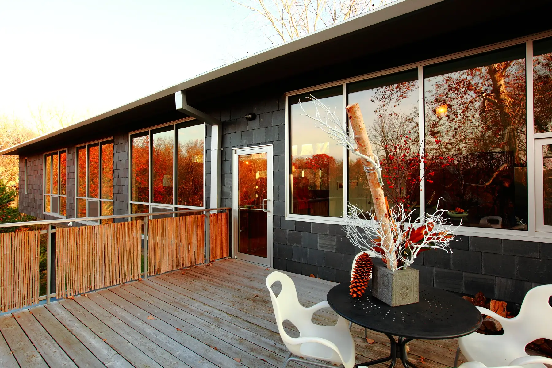 Bamboo-screened deck along the dark shingle-clad house at sunset, with large windows reflecting the autumn trees