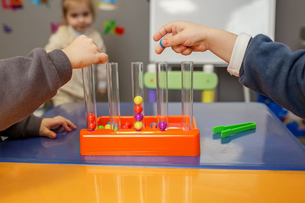Children using their hands to drop colorful beads into clear plastic test tubes during a classroom activity, demonstrating a practical example of what is play based learning.
