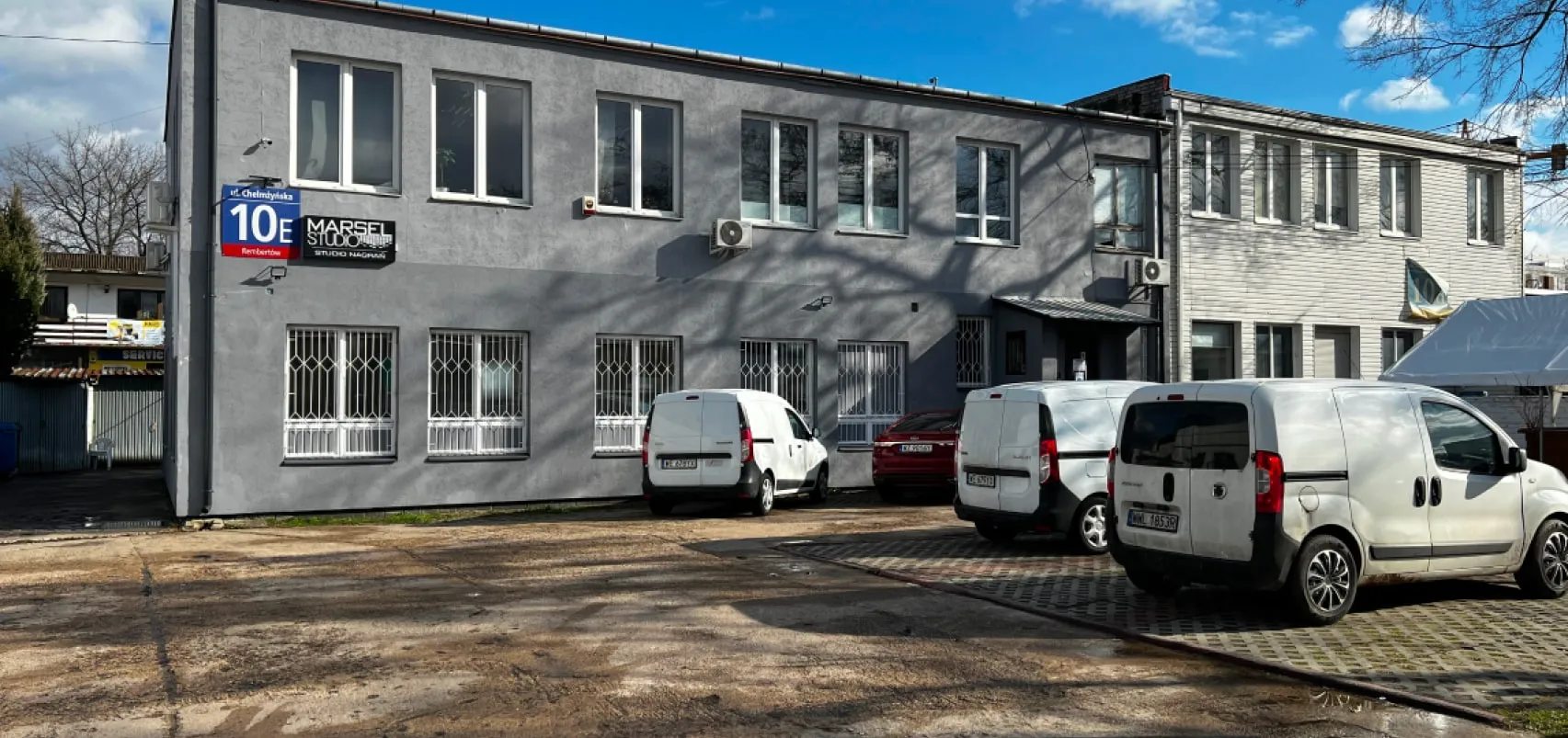 Budynek biurowy o szarych i białych elewacjach z zaparkowanymi białymi vanami i czerwonym samochodem na podwórku, znak ulicy Chełmżyńska 10E oraz tablica Marsel Studio.