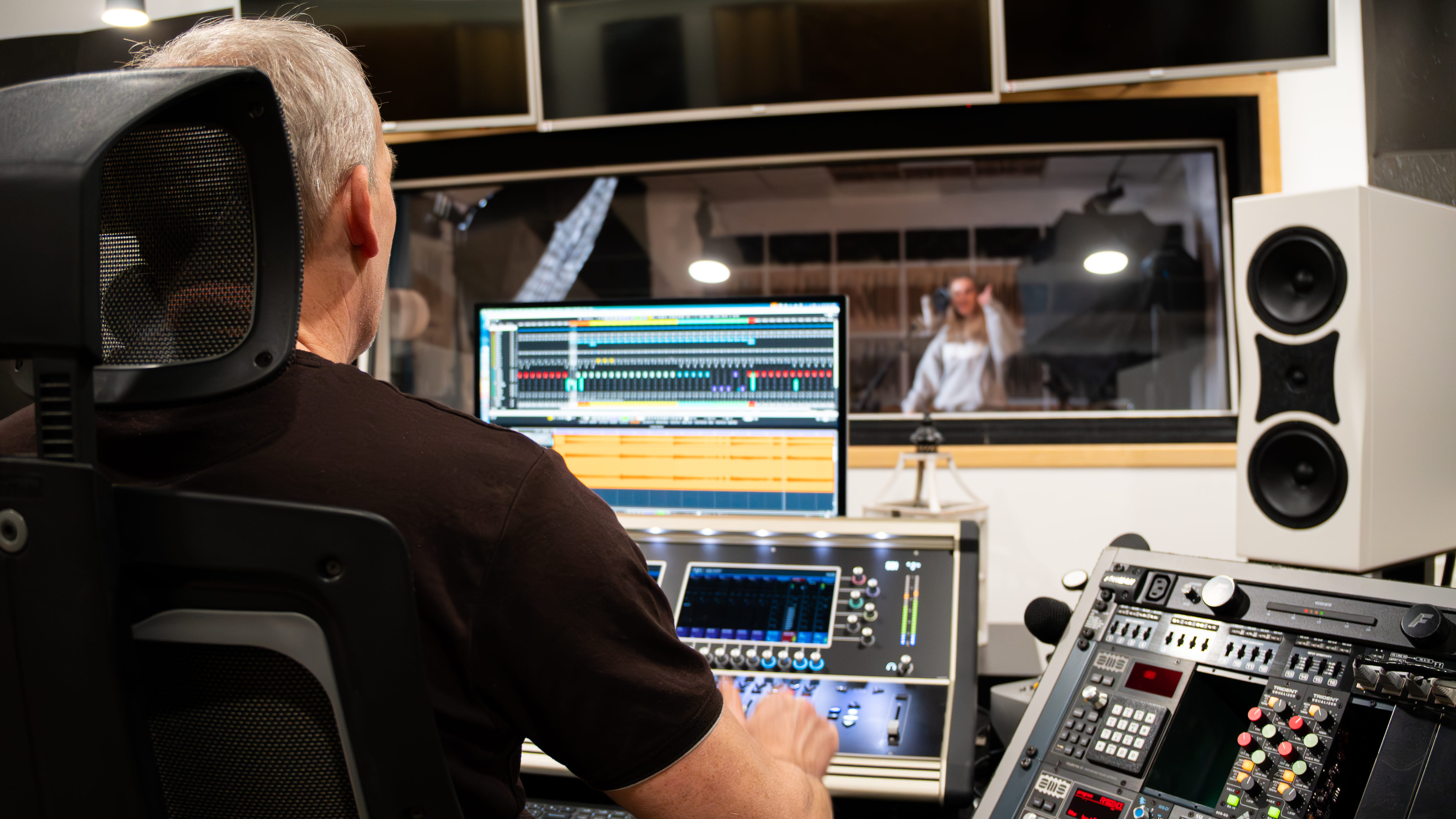 Mężczyzna siedzi przed konsolą nagraniową w studiu dźwiękowym, na ekranie widać program do edycji audio, a przez szybę znajduje się kobieta w bluzie z kapturem przed mikrofonem.