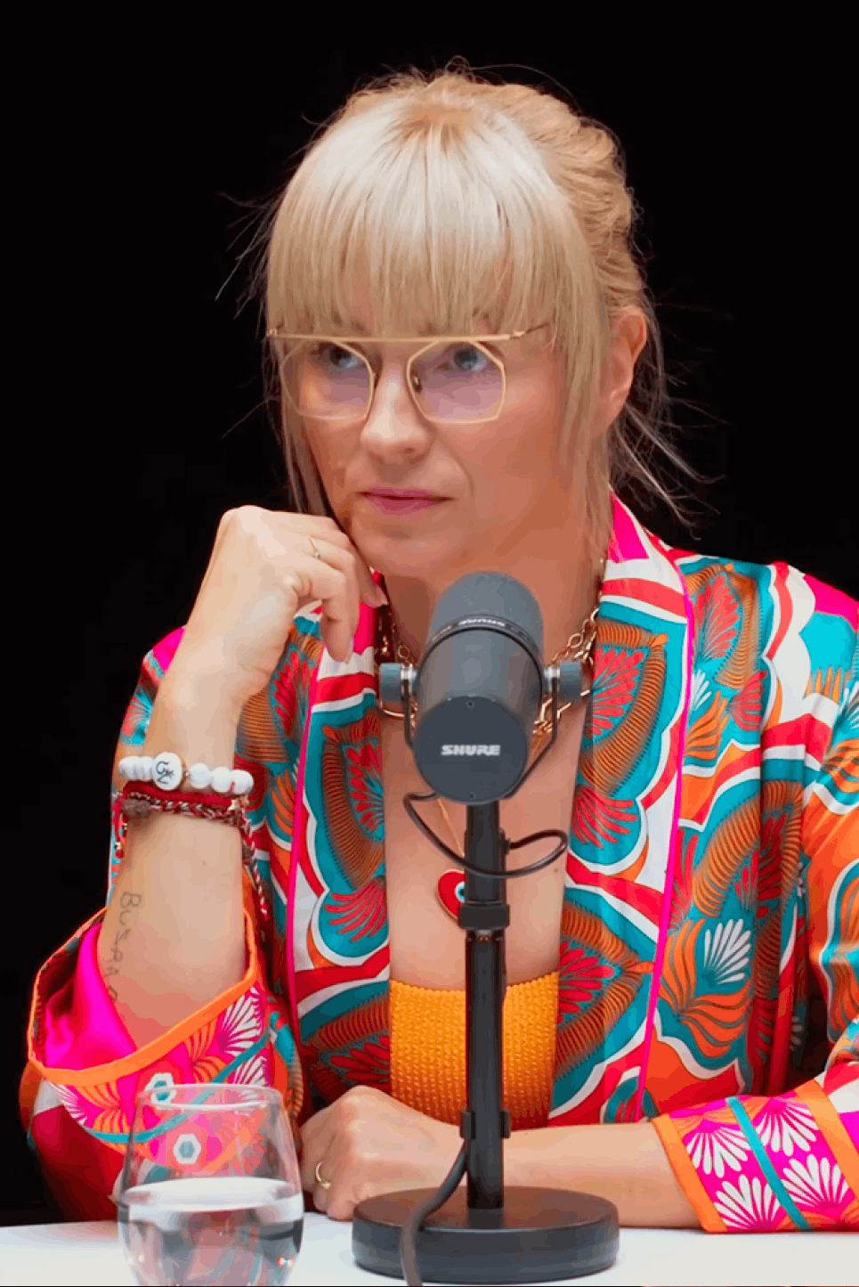 Kobieta o blond włosach i grzywce, nosząca okulary i kolorową, wzorzystą marynarkę, siedzi przy mikrofonie o nazwie Shure, na stole stoi szklanka z wodą.