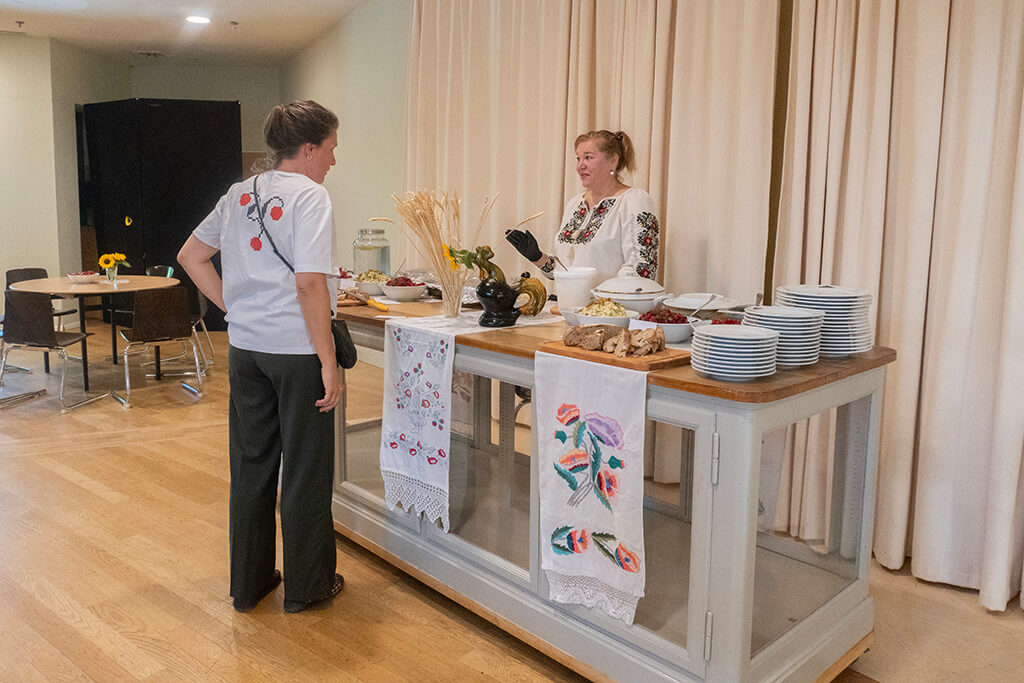 Woman in traditional embroidered blouse serving food at a buffet table decorated with embroidered cloths and ceramic dishes, talking to a guest.