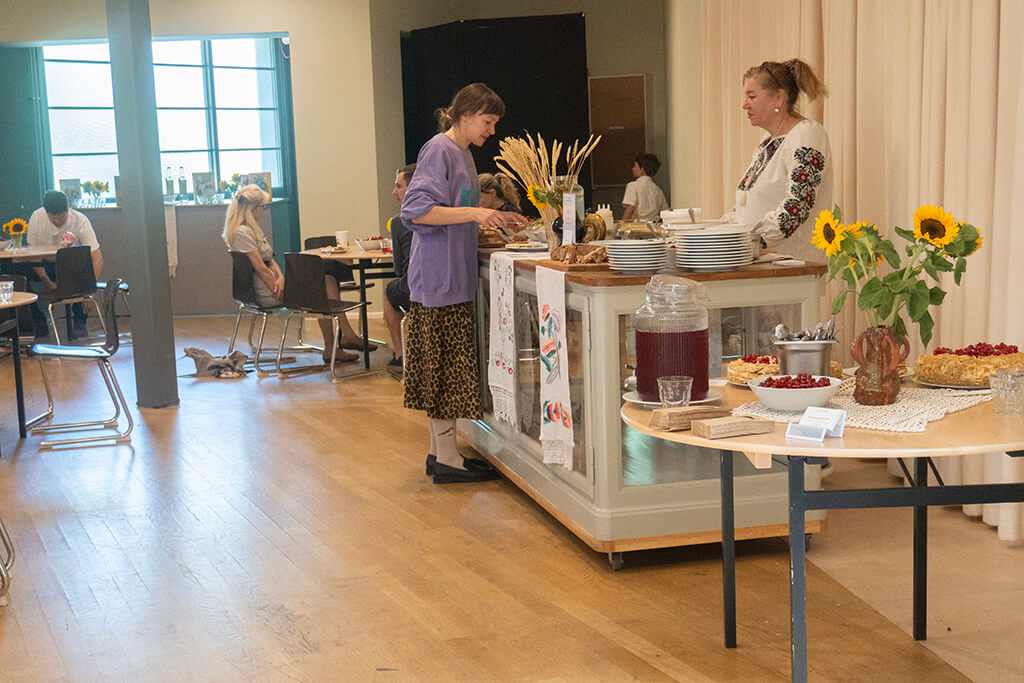 Indoor scene of a small gathering with two women near a buffet table decorated with sunflowers and desserts, and people seated at tables in the background.
