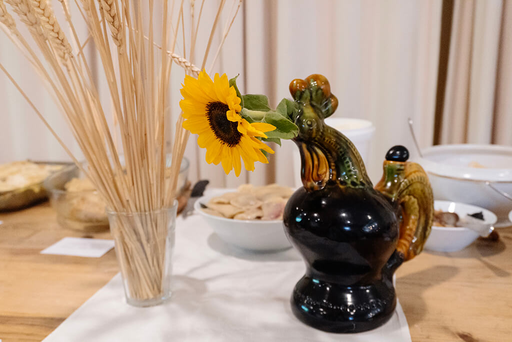 Ceramic rooster-shaped vase holding a bright yellow sunflower, next to a glass with dried wheat stalks on a table with bowls of food in the background.