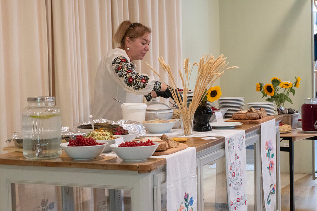 Woman serving food behind a table with bowls of berries, bread, floral decorations, and a dispenser of infused water.