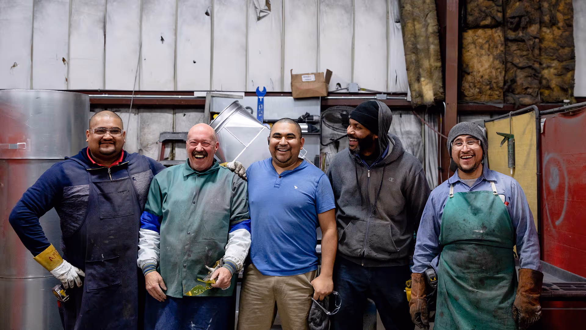 Five male workers in protective clothing smiling and standing together inside an industrial workshop.
