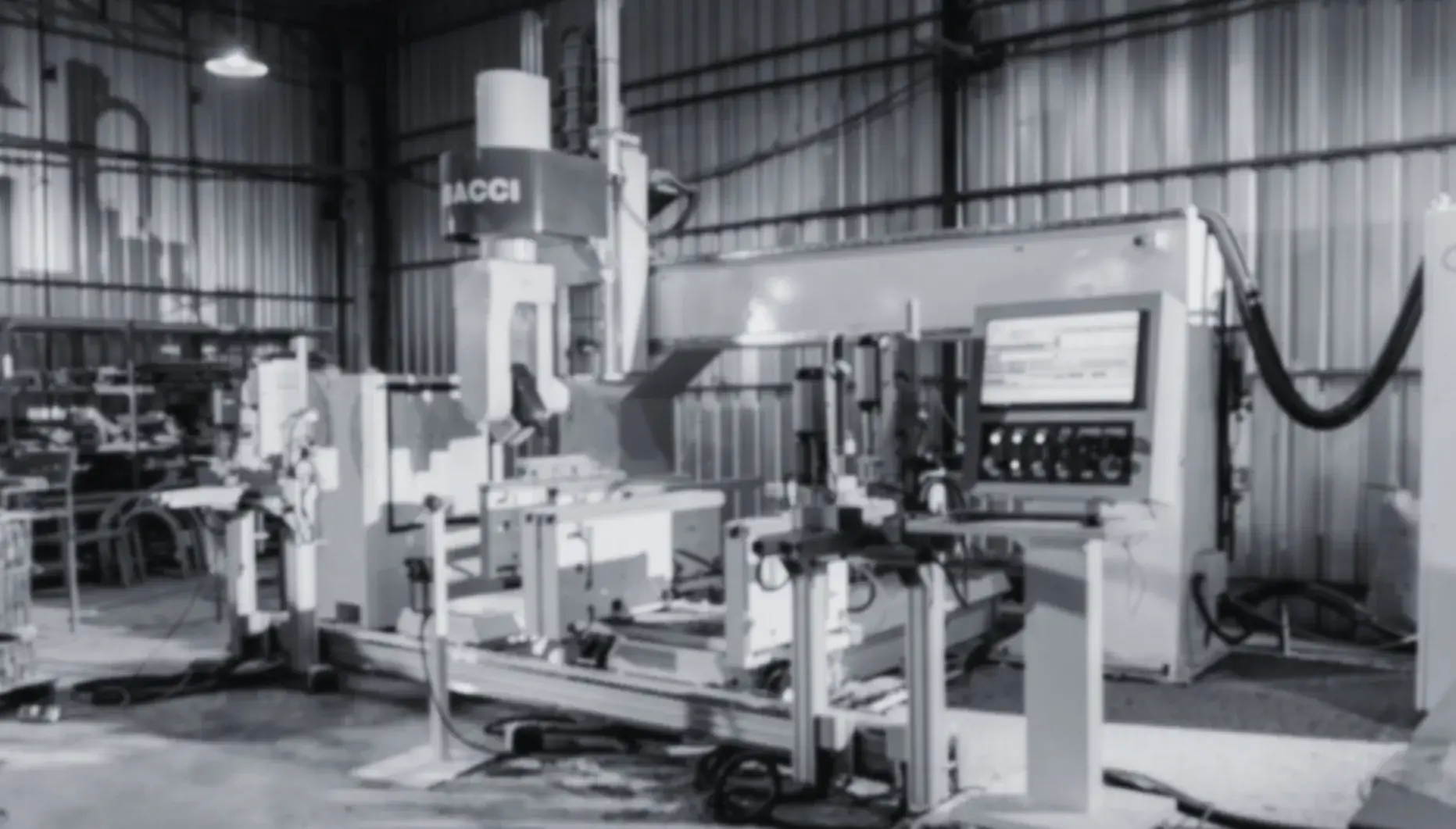 The image appears to be a black and white photograph of an industrial setting, specifically inside what looks like a large manufacturing or engineering facility.

In the center of the photo is a large, sophisticated piece of machinery with multiple components and articulated arms, suggesting it is a CNC (Computer Numerical Control) milling machine or similar type of advanced machining tool. The machine has what seems to be a spindle visible at the top center, which is consistent with milling operations. A sign on the machine reads "FACCI," which could indicate the manufacturer of the machinery. The machine stands on a sturdy base equipped with various clamps and fixtures that hold workpieces securely during operation.

To the right side of the frame, there's an electronic control cabinet with multiple screens displaying what could be programming software for the machine. Next to this panel is a keyboard, likely for inputting commands into the machine's system.

On the floor, we see cables and hoses, possibly connected to the machinery for power, coolant, or data communication. These cables are indicative of a well-equipped workspace that requires precise power and signal management.

The background reveals part of a metal structure wall and ceiling, reinforcing the industrial setting. There is also a light source overhead, casting shadows on the floor, which suggests an indoor environment with artificial lighting.

Overall, the image presents a scene of high-tech manufacturing capability, where precision engineering and automation play key roles in the production process.