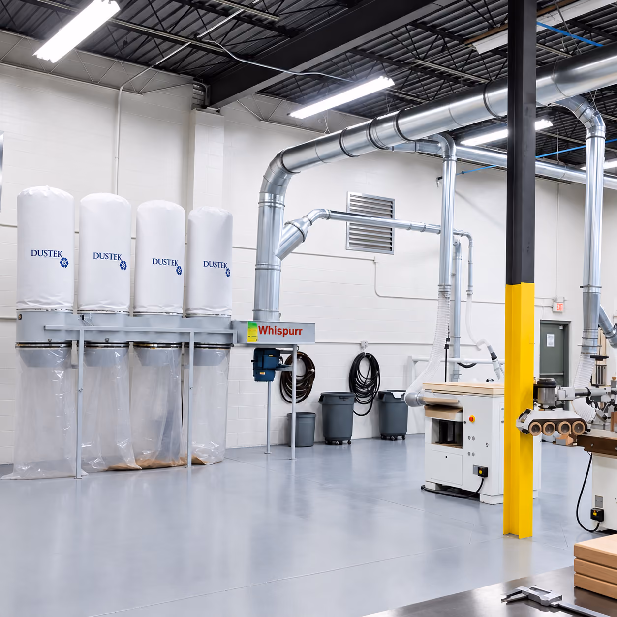 Industrial workshop dust collection system with four white DUSTEK filters and metal ductwork overhead.