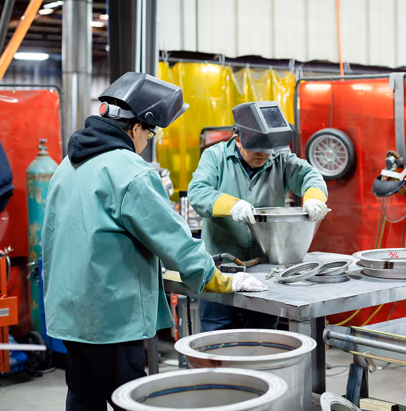 Two workers in protective gear assembling metal duct parts in an industrial workshop.